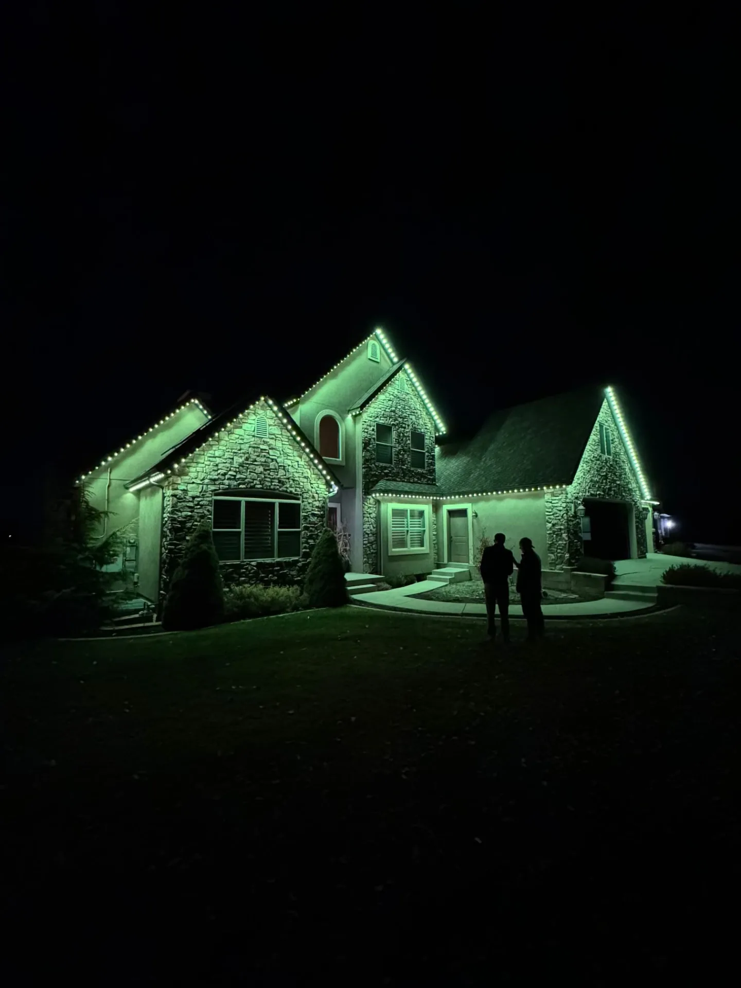 Stone home lit in green with homeowners admiring it